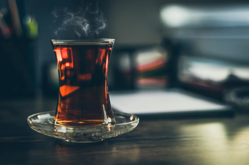 Close-up photograph of steaming black tea in a clear glass cup, showing the dark amber liquid with visible steam rising, shot by Hasan Albari